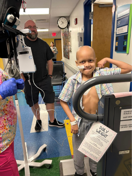 A child with a nasal tube smiles and flexes his arm near a hospital scale. An adult and a nurse stand nearby in a hospital hallway. A sign reads Daily Weight..
