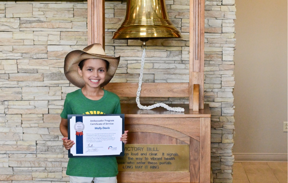 A smiling child in a cowboy hat holds a certificate and stands next to a large golden bell mounted on a wooden base against a stone wall.