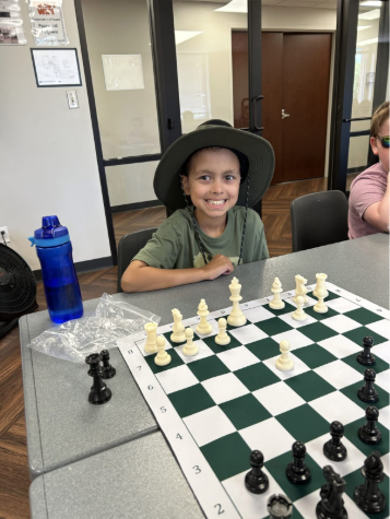 A smiling child wearing a wide-brimmed hat sits at a table with a chessboard, ready to play. A blue water bottle and chess pieces are on the table.