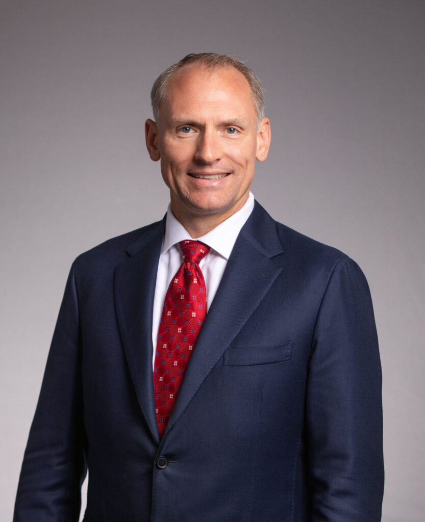 A man in a navy suit, white shirt and red patterned tie stands smiling against a plain gray background.