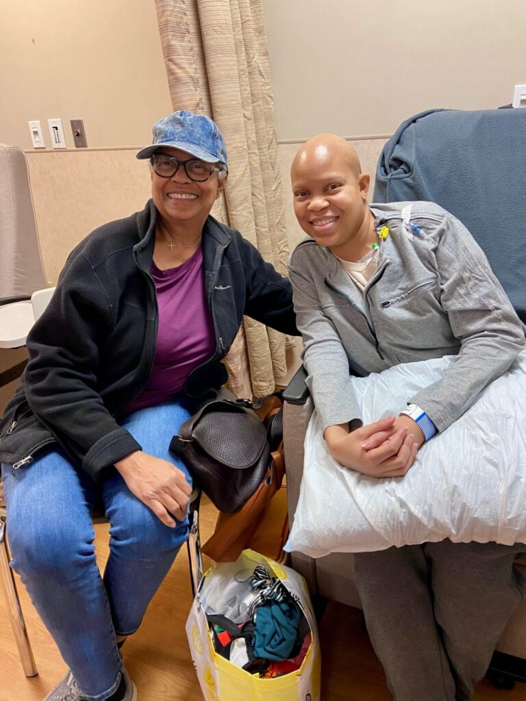 Two women sitting side-by-side in a an infusion room, both smiling at the camera.
