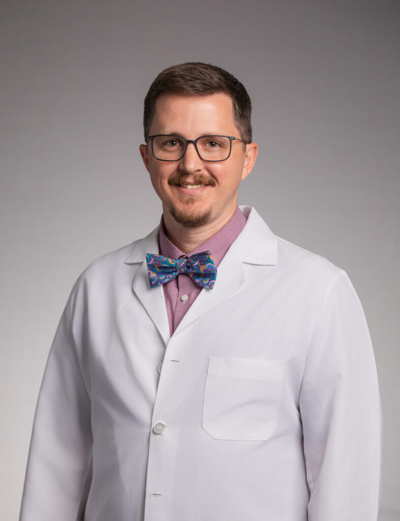 A man with a mustache and glasses wearing a white lab coat over a red collared shirt and a colorful patterned bowtie.
