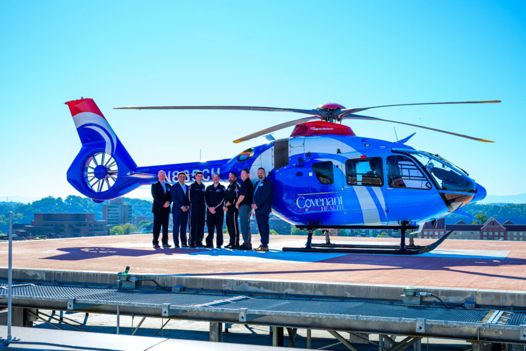 Seven people standing in a row in front of a blue Covenant Health medical helicopter on a rooftop helipad.