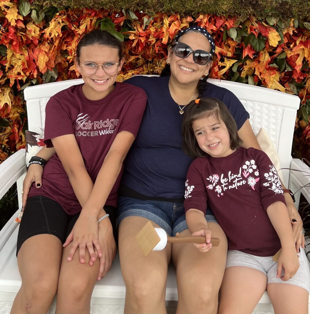 Three people sit on a white bench before a wall of fall leaves, smiling at the camera. The youngest, in a maroon shirt with white flowers, holds a wooden spatula—a cheerful touch to their aquablation celebration.