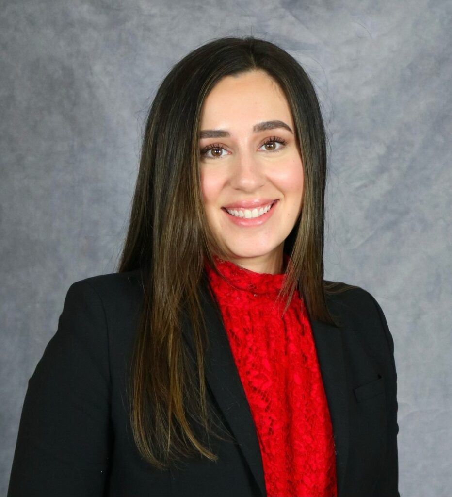 A woman with long brown hair, wearing a black blazer over a red lace top, smiling against a gray background.