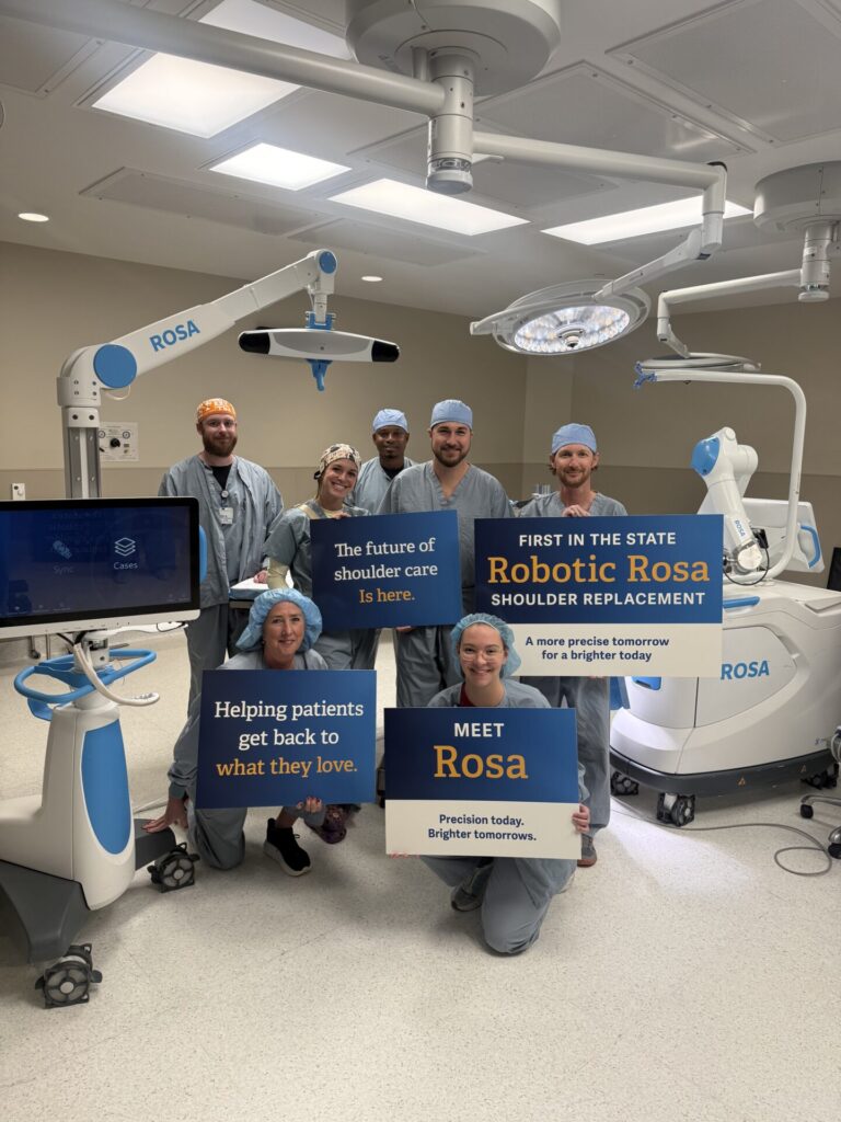 A surgical team in a hospital operating room holds signs promoting Robotic Rosa shoulder replacement technology.