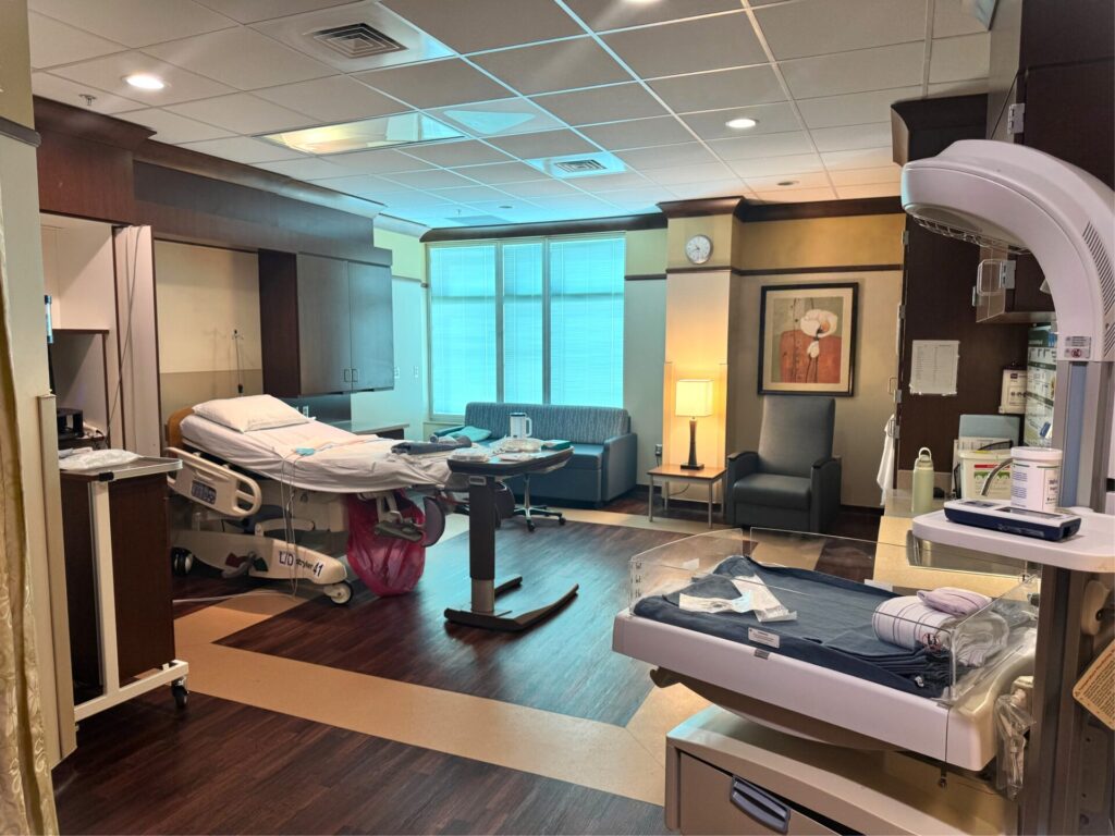 Hospital room with an adjustable bed, an infant bassinet in the foreground, and a seating area with a sofa and armchair.