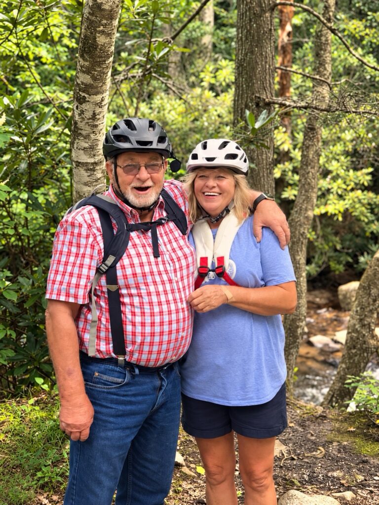 A smiling man in a plaid shirt and a woman in a blue t-shirt standing together in a forest, both wearing bicycle helmets.