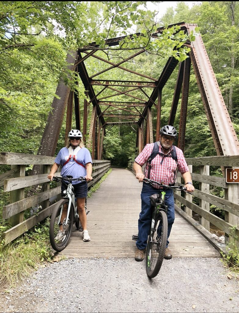 Two people wearing helmets on mountain bikes standing at the entrance of a weathered metal truss bridge.
