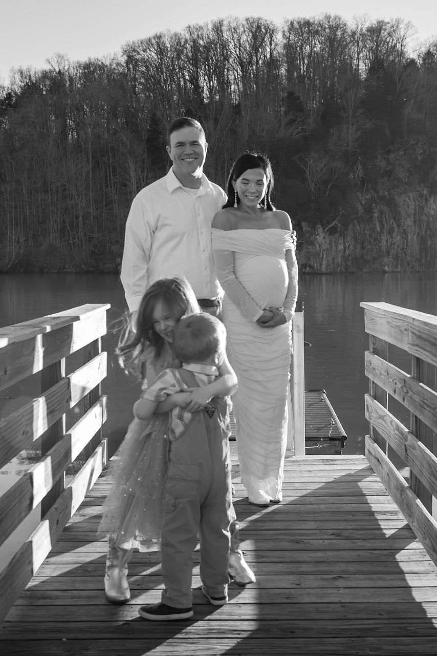 A family of four standing on a wooden pier by a lake, with parents standing behind two young children who are hugging.