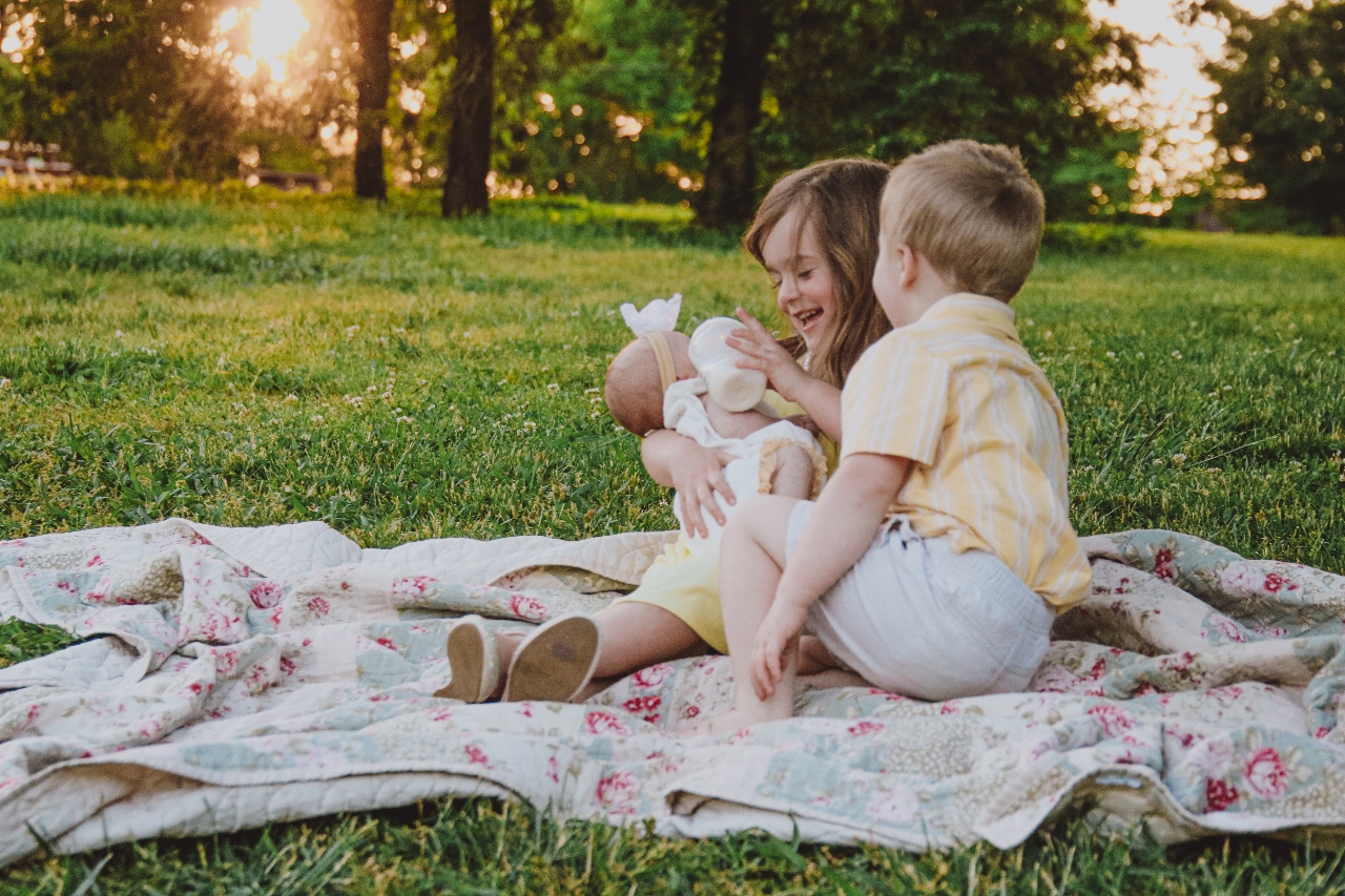 A young girl and boy sit on a quilt in a grassy field, with the girl feeding a baby from a bottle.