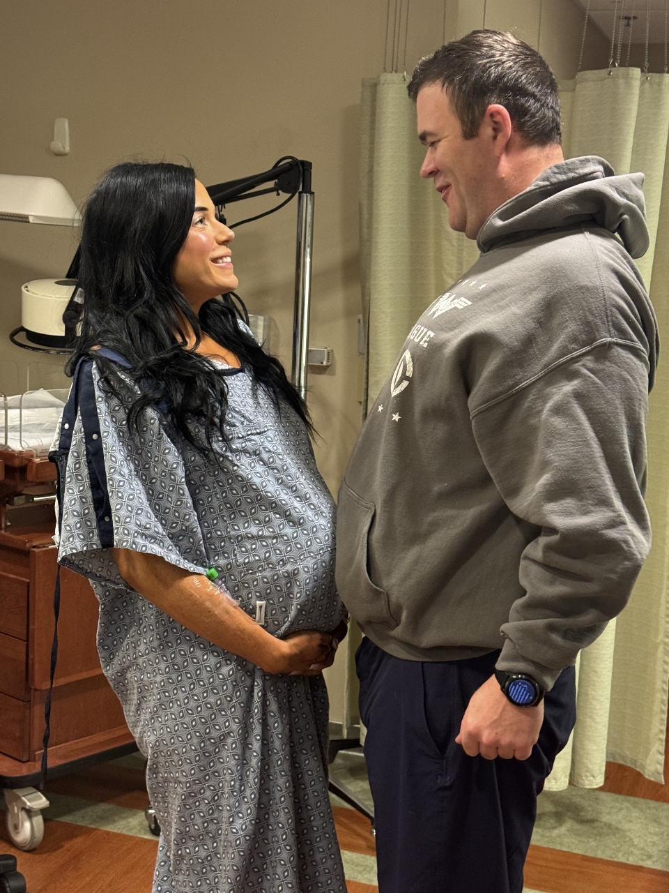 A pregnant woman in a hospital gown and a man in a hooded sweatshirt stand facing each other and smiling.