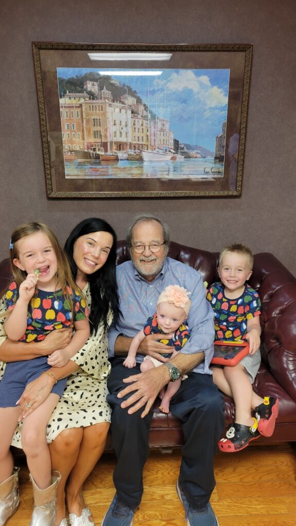 A multi-generational family sitting on a leather sofa, with a painting of a coastal town on the wall behind them.
