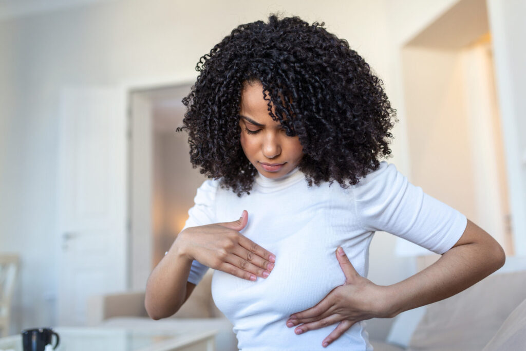 A woman with curly hair in a white shirt examines her breast with her hand, appearing to perform a self-breast check at home.