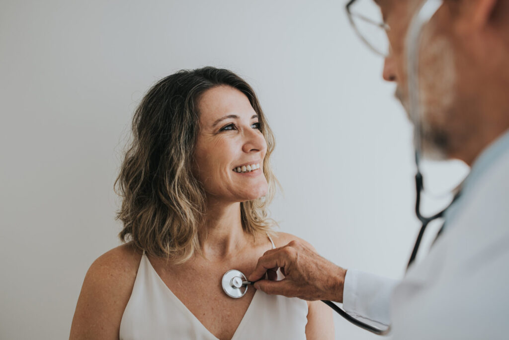 A doctor listens to a smiling patients chest with a stethoscope.