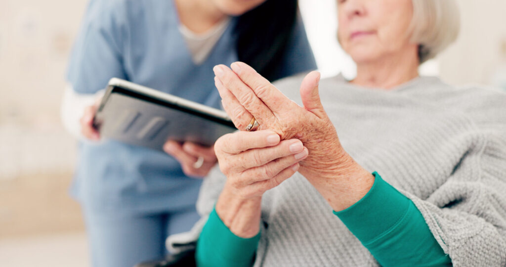 An elderly woman sitting and holding her painful hand while a healthcare professional in scrubs stands nearby with a clipboard.