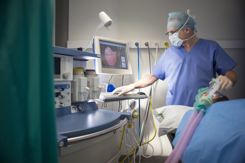 A surgeon in blue scrubs, mask, and cap monitors an ultrasound display while attending to an anesthetized patient.